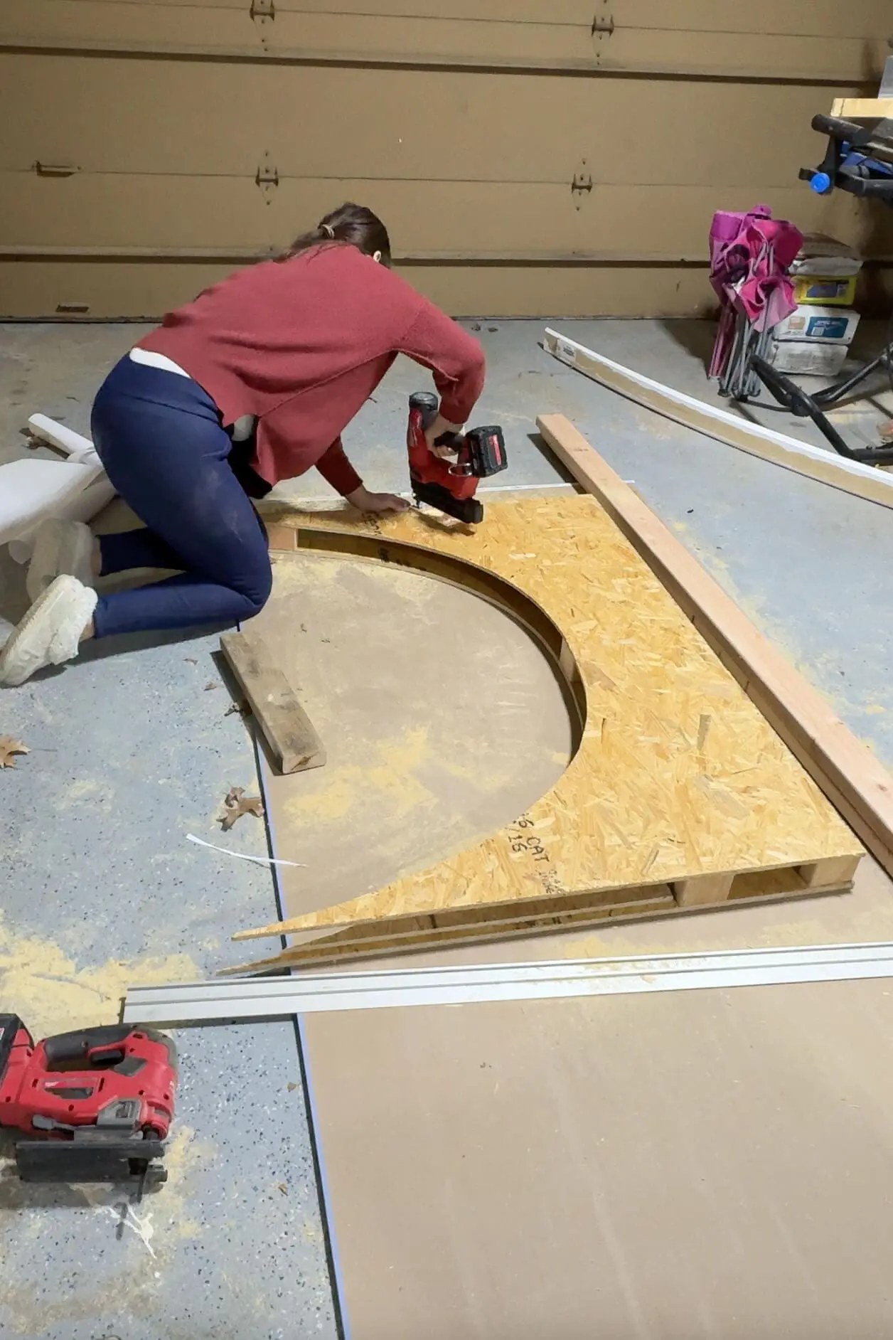 A woman kneels and uses a red brad nailer to build a plywood arch. The garage floor shows visible sawdust and additional construction materials.